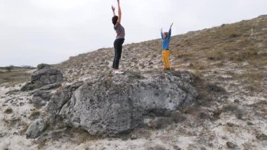 Bir anne ve çocuk engebeli bir arazide kayalar üzerinde esneme egzersizleri yapıyorlar. Çocuk onu takip ederken anne kollarını yukarı kaldırır. Doğada aktif bir spor yapmayı seviyorlar..