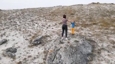 Bir anne ve çocuk engebeli bir arazide kayalar üzerinde esneme egzersizleri yapıyorlar. Çocuk onu takip ederken anne kollarını yukarı kaldırır. Doğada aktif bir spor yapmayı seviyorlar..