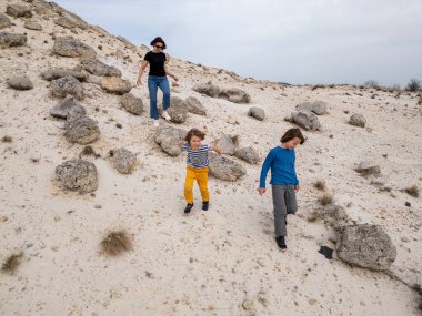 Bir anne ve iki çocuk kayalık bir açık araziyi keşfediyorlar. Arazi engebelidir. Arka planda seyrek bitki örtüsü ve uzak tepeler vardır.