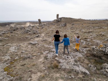 Bir anne ve iki çocuk kayalık bir açık araziyi keşfediyorlar. Arazi engebelidir. Arka planda seyrek bitki örtüsü ve uzak tepeler vardır.