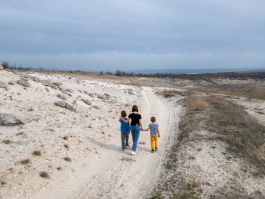 Bir anne ve iki çocuk kayalık bir açık araziyi keşfediyorlar. Arazi engebelidir. Arka planda seyrek bitki örtüsü ve uzak tepeler vardır.
