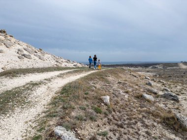 Bir anne ve iki çocuk kayalık bir açık araziyi keşfediyorlar. Arazi engebelidir. Arka planda seyrek bitki örtüsü ve uzak tepeler vardır.