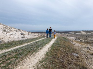 Bir anne ve iki çocuk kayalık bir açık araziyi keşfediyorlar. Arazi engebelidir. Arka planda seyrek bitki örtüsü ve uzak tepeler vardır.