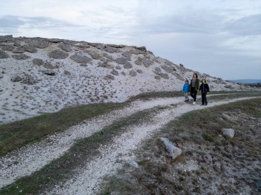 Kuru, açık bir arazide, kumlu bir patika boyunca iki çocukla yürüyen bir kadın. Dağılmış kayalar ve bulutlu bir gökyüzünün altında seyrek bitki örtüsü..
