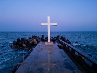 Alacakaranlıkta deniz kıyısındaki beton bir iskelede aydınlanmış bir haç heykeli, açık bir akşam gökyüzünün altında, tetrapodlara çarpan dalgalar..
