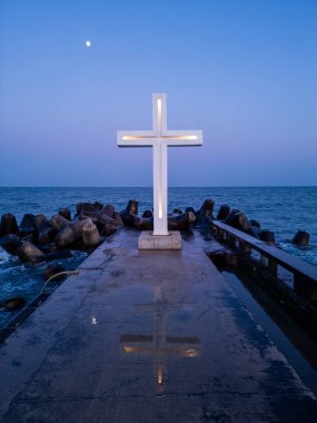 Alacakaranlıkta deniz kıyısındaki beton bir iskelede aydınlanmış bir haç heykeli, açık bir akşam gökyüzünün altında, tetrapodlara çarpan dalgalar..