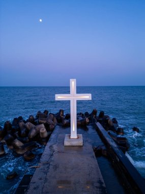 Alacakaranlıkta deniz kıyısındaki beton bir iskelede aydınlanmış bir haç heykeli, açık bir akşam gökyüzünün altında, tetrapodlara çarpan dalgalar..