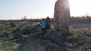 Bulutlu bir günde kayalık bir araziyi keşfeden kadın ve iki çocuk, kuru ve doğal bir ortamda yıpranmış taş oluşumlarının arasında yürüyorlar..