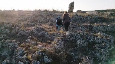 Bulutlu bir günde kayalık bir araziyi keşfeden kadın ve iki çocuk, kuru ve doğal bir ortamda yıpranmış taş oluşumlarının arasında yürüyorlar..