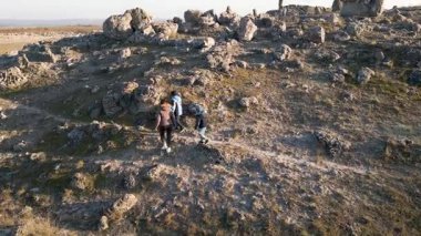 Bulutlu bir günde kayalık bir araziyi keşfeden kadın ve iki çocuk, kuru ve doğal bir ortamda yıpranmış taş oluşumlarının arasında yürüyorlar..