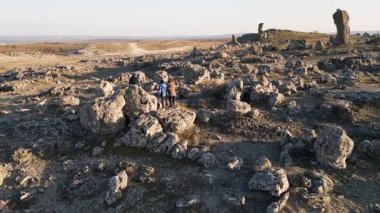 Bulutlu bir günde kayalık bir araziyi keşfeden kadın ve iki çocuk, kuru ve doğal bir ortamda yıpranmış taş oluşumlarının arasında yürüyorlar..