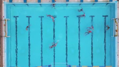 Bir su topu maçının hava görüntüsü devam ediyor, oyuncular açık mavi bir havuzda yüzüyor ve yarışıyorlar, her iki uçta da görünür hedefler var.