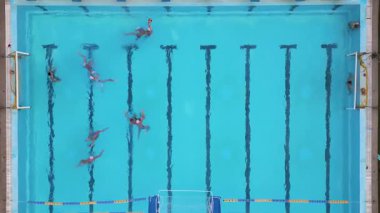 Bir su topu maçının hava görüntüsü devam ediyor, oyuncular açık mavi bir havuzda yüzüyor ve yarışıyorlar, her iki uçta da görünür hedefler var.