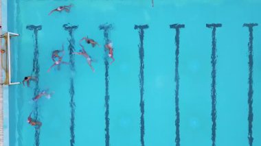 Bir su topu maçının hava görüntüsü devam ediyor, oyuncular açık mavi bir havuzda yüzüyor ve yarışıyorlar, her iki uçta da görünür hedefler var.