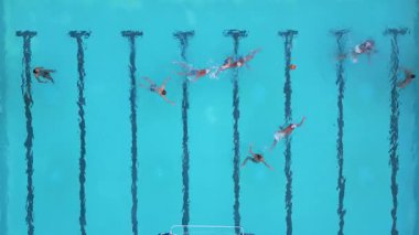 Bir su topu maçının hava görüntüsü devam ediyor, oyuncular açık mavi bir havuzda yüzüyor ve yarışıyorlar, her iki uçta da görünür hedefler var.