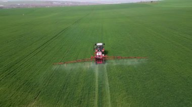 İlkbaharda geniş yeşil bir alana gübre ya da böcek ilacı püskürten traktör modern tarım teknolojisi ve mahsul bakımını gösteriyor..