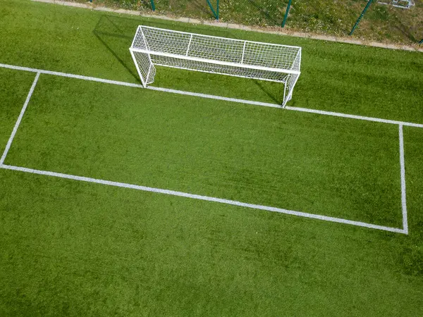 Beyaz çizgileri olan yeşil bir suni çim sahasındaki futbol kalesinin havadan görünüşü. Canlı karşıtlık ve geometrik düzen spor atmosferini ve rekabetçi ruhu vurguluyor.
