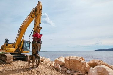 Hidrolik pençeli sarı kazıcı parçalı bulutlu bir gökyüzünün altında deniz kenarındaki kayalık bir sahil inşaatında çalışıyor..