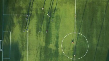 Yeşil bir sahada, öğleden sonra güneşinde oyuncuların oynadığı uzun gölgeli bir gençlik futbol antrenmanının havadan görüntüsü..