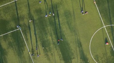 Yeşil bir sahada, öğleden sonra güneşinde oyuncuların oynadığı uzun gölgeli bir gençlik futbol antrenmanının havadan görüntüsü..