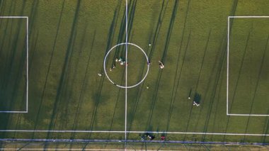 Yeşil bir sahada, öğleden sonra güneşinde oyuncuların oynadığı uzun gölgeli bir gençlik futbol antrenmanının havadan görüntüsü..
