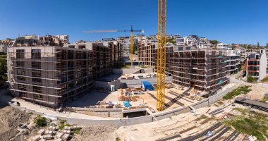 Şehir ortamında açık mavi gökyüzü altında iskelesi, vinçleri ve bitmemiş binaları olan modern bir yerleşim alanının panoramik görüntüsü.