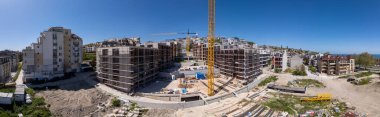 Şehir ortamında açık mavi gökyüzü altında iskelesi, vinçleri ve bitmemiş binaları olan modern bir yerleşim alanının panoramik görüntüsü.
