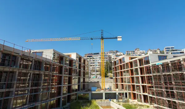 Şehir ortamında açık mavi gökyüzü altında iskelesi, vinçleri ve bitmemiş binaları olan modern bir yerleşim alanının panoramik görüntüsü.