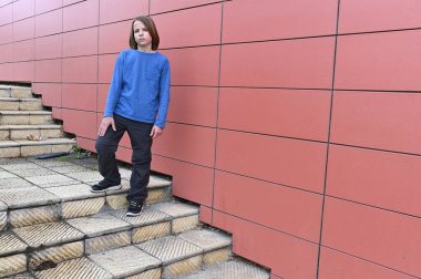 Çocuk, mavi bir gömlek ve siyah pantolon giymiş, dokulu bir kaldırımda, büyük kırmızı fayanslı bir duvara karşı duruyor, sakin ve düşünceli görünüyor şehir ortamında.