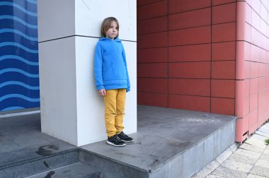 Mavi kapüşonlu ve sarı pantolonlu genç beyaz bir sütunun yanında beton basamaklarda duruyor ve arka planda dalga deseni olan kırmızı fayanslı bir duvar..