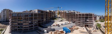 Şehir ortamında açık mavi gökyüzü altında iskelesi, vinçleri ve bitmemiş binaları olan modern bir yerleşim alanının panoramik görüntüsü.