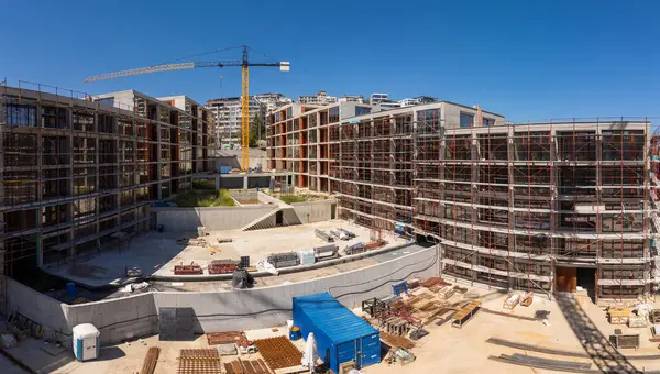 Şehir ortamında açık mavi gökyüzü altında iskelesi, vinçleri ve bitmemiş binaları olan modern bir yerleşim alanının panoramik görüntüsü.