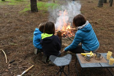 Anne ve iki çocuk ormanda bir kamp ateşinin yanında oturur, küçük bir masada yemek yer ve dinlenirler. Çam ağaçları ve doğa ile çevrili sıcak bir açık hava yemeğinden ve aile zamanından hoşlanırlar..