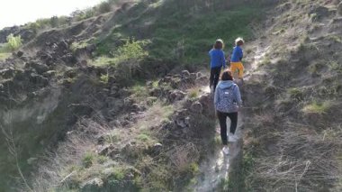 Anne ve iki çocuk, kayalar ve bahar bitkileriyle çevrili dar bir patika boyunca yokuş yukarı yürüyorlar. Güneşli bir günde, doğal bir ortamda huzurlu bir açık hava macerası..
