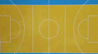 Renkli bir açık hava basketbol sahasının havadan görünüşü. Canlı mavi ve sarı işaretler, iki halka ve simetrik yerleşim. Doğrudan yukarıdan ateş edilmiş.