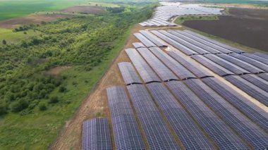 Yeşil alanlar ve elektrik hatları ile çevrili kırsal bir alanda sıra sıra fotovoltaik paneller kurulmuş büyük bir güneş enerjisi çiftliğinin havadan görünüşü. Yenilenebilir enerji kavramı.