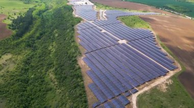Yeşil alanlar ve elektrik hatları ile çevrili kırsal bir alanda sıra sıra fotovoltaik paneller kurulmuş büyük bir güneş enerjisi çiftliğinin havadan görünüşü. Yenilenebilir enerji kavramı.