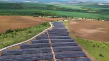 Yeşil alanlar ve elektrik hatları ile çevrili kırsal bir alanda sıra sıra fotovoltaik paneller kurulmuş büyük bir güneş enerjisi çiftliğinin havadan görünüşü. Yenilenebilir enerji kavramı.