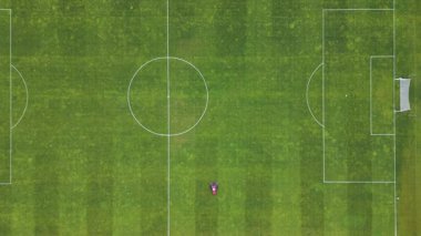 Futbol sahasında çim biçen kırmızı bir çim biçme makinesinin havadan görünüşü çizgili desenler yaratıyor. Yeşil yemyeşil bir zeminde spor sahasının bakımı devam ediyor.