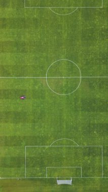 Futbol sahasında çim biçen kırmızı bir çim biçme makinesinin havadan görünüşü çizgili desenler yaratıyor. Yeşil yemyeşil bir zeminde spor sahasının bakımı devam ediyor.