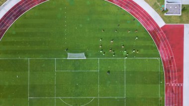 Yeşil bir futbol sahasında oyuncuların antrenman yaptığı bir futbol antrenmanının hava görüntüsü. Koşu pisti ve kale direği görünüyor. Spor pratiği ve takım çalışması konsepti.