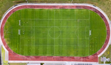 Kırmızı koşu pisti ile çevrili, birden fazla eğitim işareti olan büyük bir futbol sahasının hava görüntüsü. Sporcular tatbikat yaparken görülüyor. Yukarıdan spor ve spor kompleksi.
