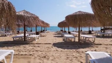 Güneşli bir günde sakin mavi denize bakan saman şemsiyeli ve beyaz güneşli kumsal. Huzurlu yaz sahnesi tatil, dinlenme ve seyahat promosyonları için ideal..