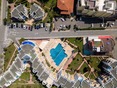 Yüzme havuzu, güneşli salonlar, manzaralı bahçeler ve çevre binaları olan modern bir konut kompleksinin tepeden tırnağa görüntüsü. Güneşli bir gün, geometrik yerleşim ve park edilmiş arabalar görünür..
