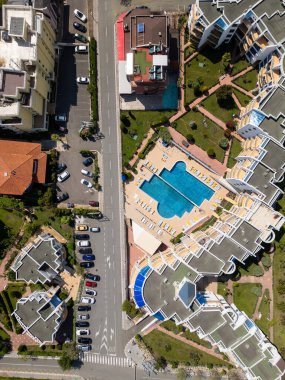 Yüzme havuzu, güneşli salonlar, manzaralı bahçeler ve çevre binaları olan modern bir konut kompleksinin tepeden tırnağa görüntüsü. Güneşli bir gün, geometrik yerleşim ve park edilmiş arabalar görünür..