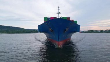 Sakin sularda yüzen renkli konteynırlarla dolu büyük mavi bir kargo gemisinin önü. Martıları ve kıyı arkaplanı olan sanayi deniz manzarası.