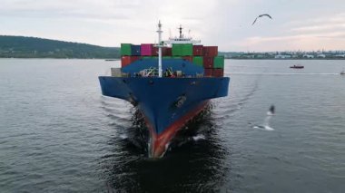 Sahil kenarında sakin sularda yüzen renkli konteynırlar taşıyan büyük mavi bir kargo gemisinin ön görüntüsü. Arka planda martılar ve liman altyapısı olan sanayi sahnesi.