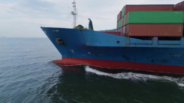 Sakin sularda giden konteynırlarla dolu büyük mavi bir kargo gemisinin yan görüntüsü. Endüstriyel deniz taşıma sahnesi görünür çapa ve pruva dalgalarıyla.