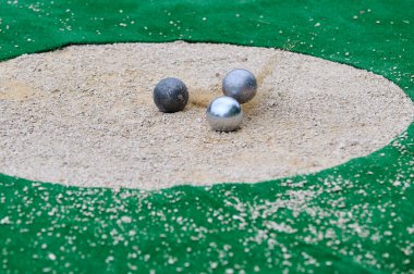 Petanque hassas atış müsabakası. Metal toplar kumda hedefe çarpıyor. Çarpışma anında toz bulutu uçuyor. Doğruluk ve dinamik oyuna odaklan.