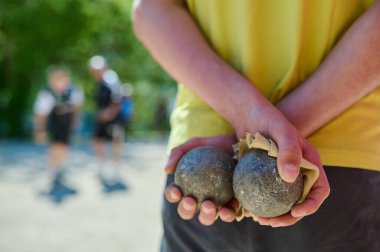 Maç sırasında arkalarında iki metal top ve bir bez tutan Petanque oyuncusu. Güneşli bir günde arka planda bulanık oyuncuların olduğu ellere ve ekipmanlara odaklan.